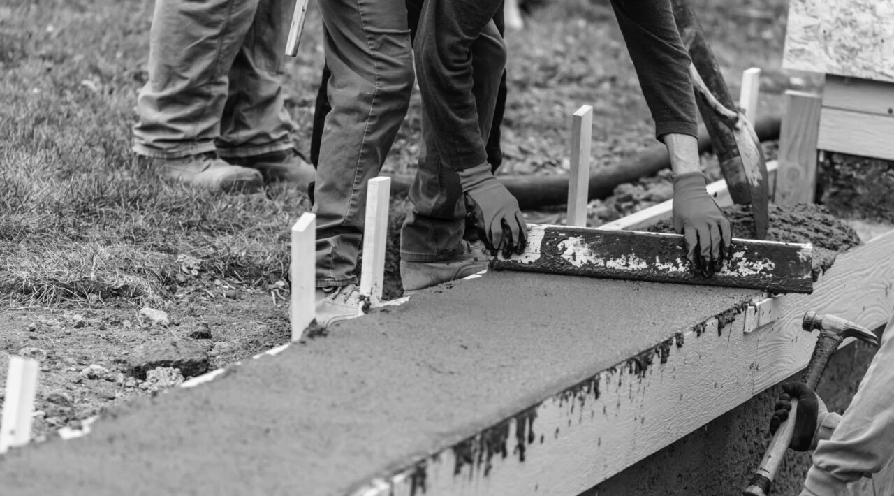 Construction Workers Pouring And Leveling Wet Cement Into Wood Framing.